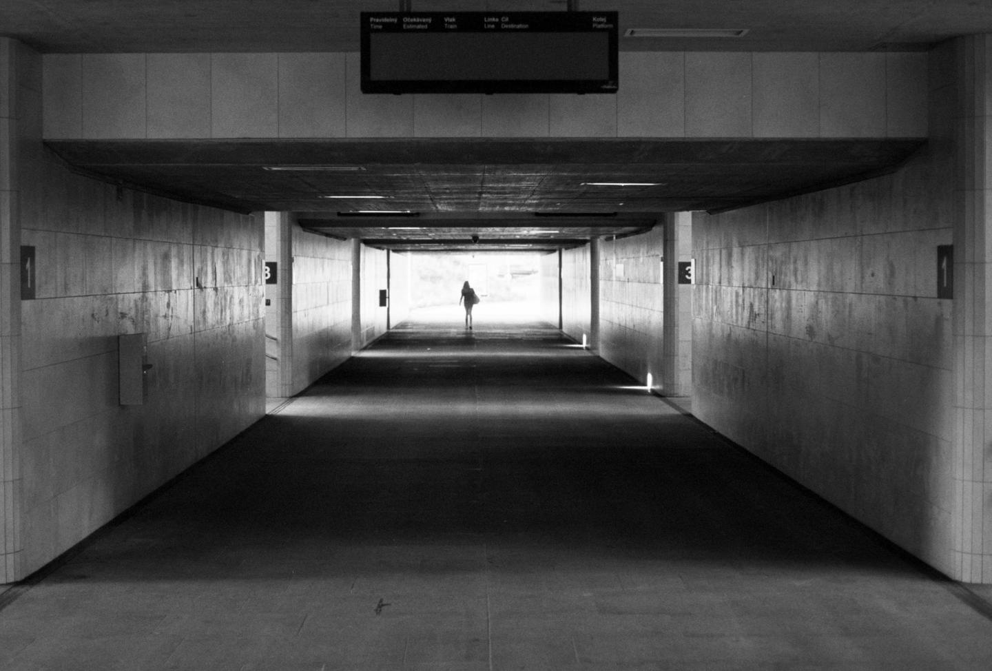 A lone figure walks towards a bright light at the end of a long, dark tunnel.