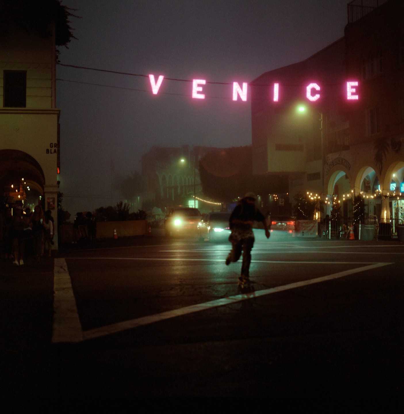 A person on a skateboard crosses a street at night under a neon "VENICE" sign.
