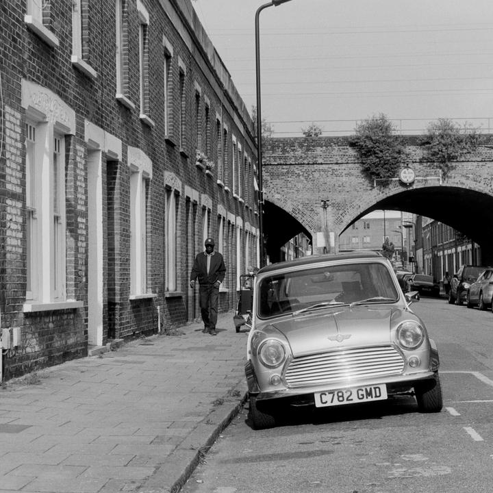 A man walks along a sidewalk next to a parked car and brick buildings under a bridge.
