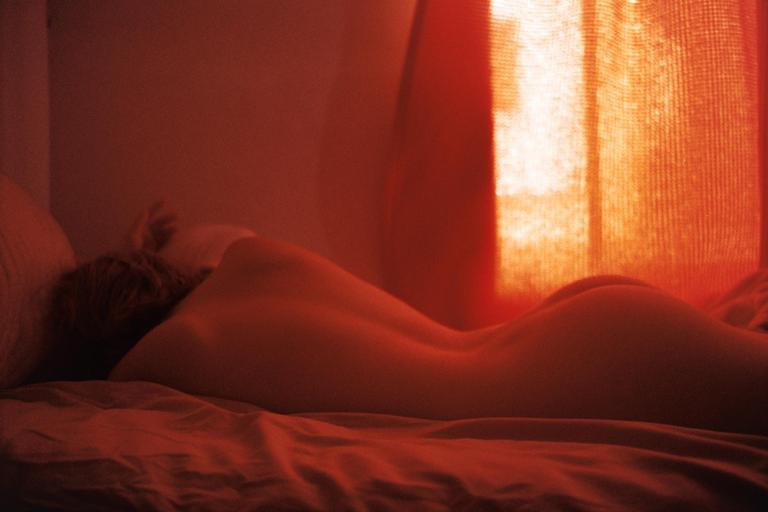A woman lies on her stomach in a bed with red light coming through a window.