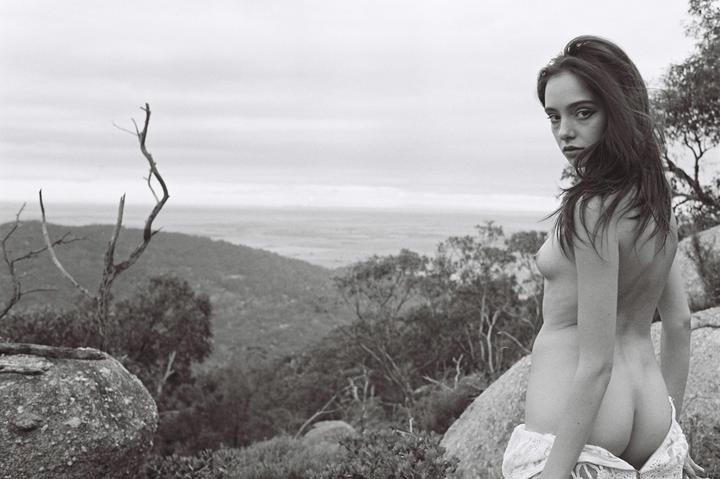 A woman stands topless on a rocky hillside overlooking a valley.