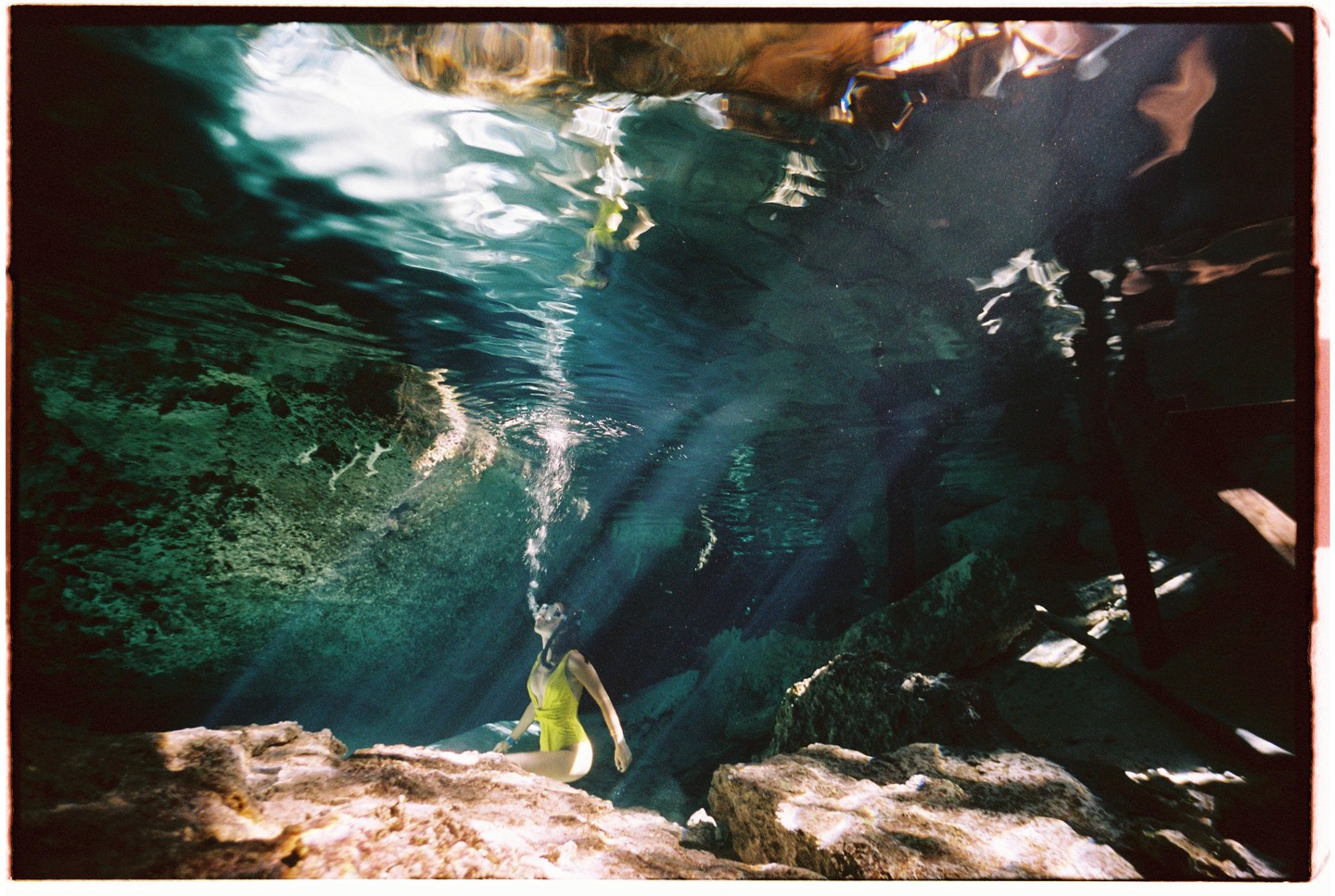 A woman in a yellow swimsuit is underwater, exhaling bubbles, with light rays shining through the water.