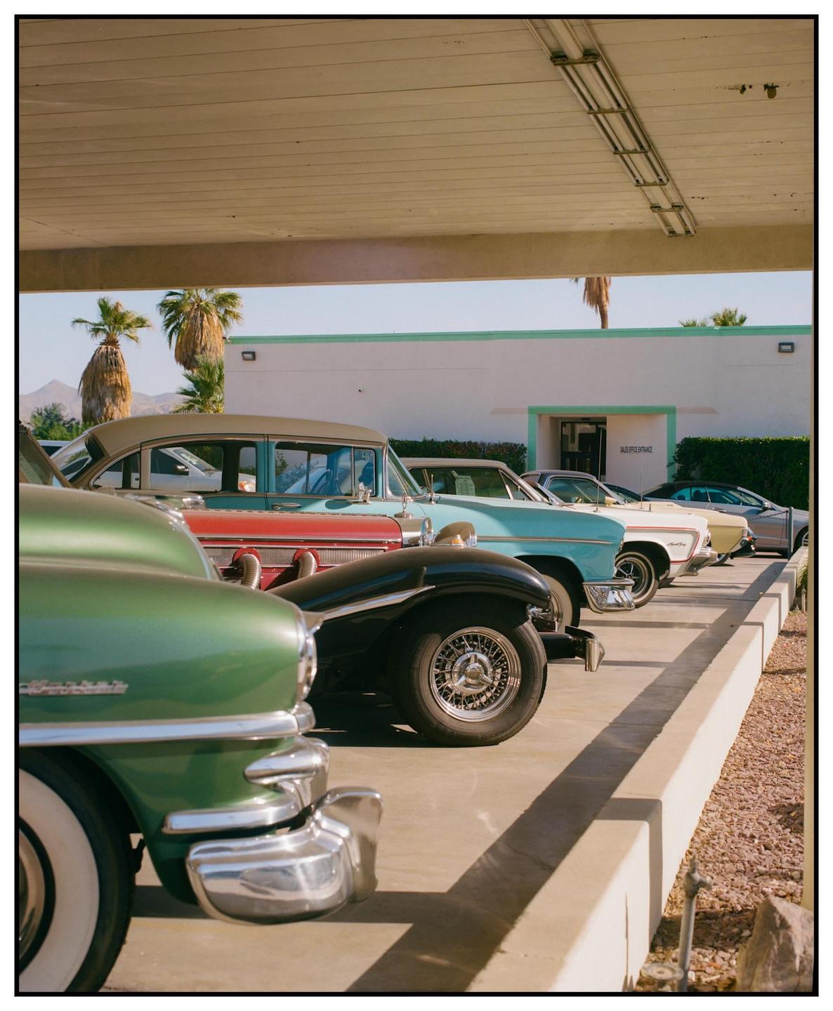 A line of vintage cars is parked under a covered structure with palm trees and a building in the background.