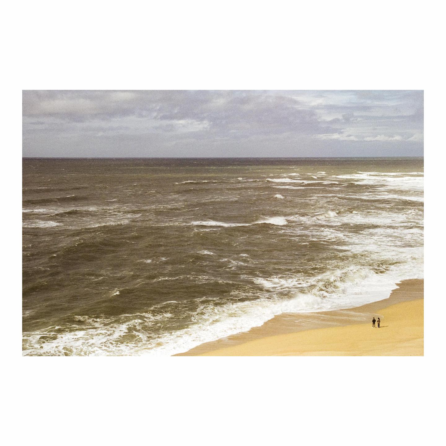 Two people stand on a sandy beach next to a rough ocean under a cloudy sky.