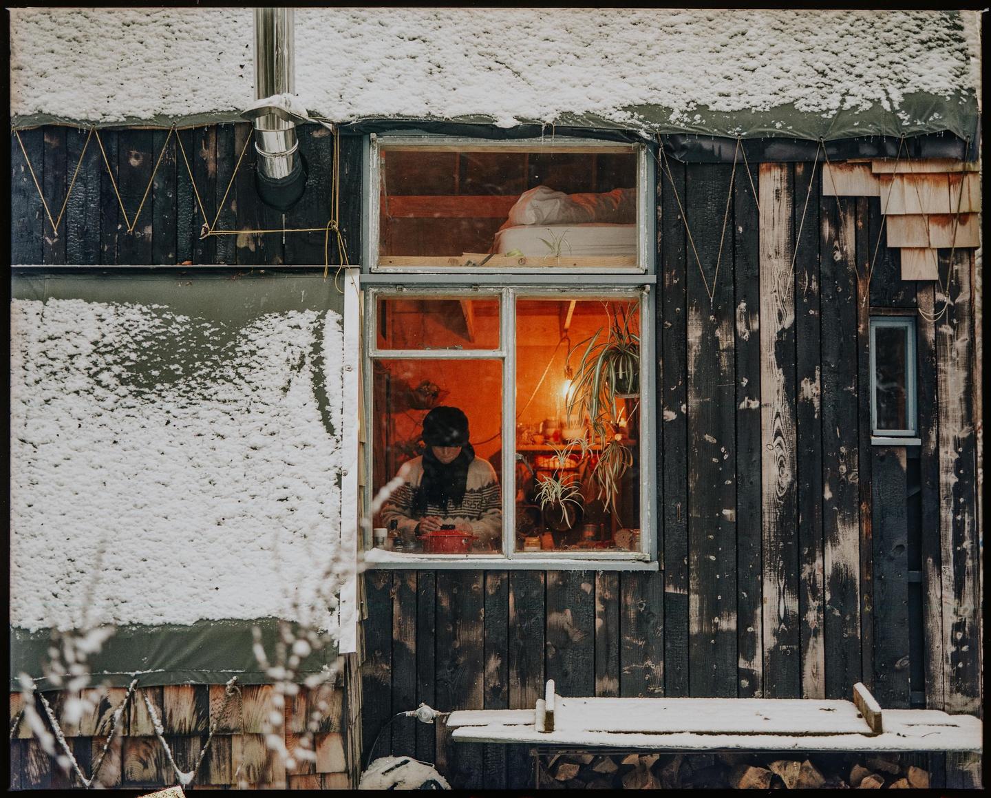 A woman sits inside a cabin with snow on the roof and ground outside.