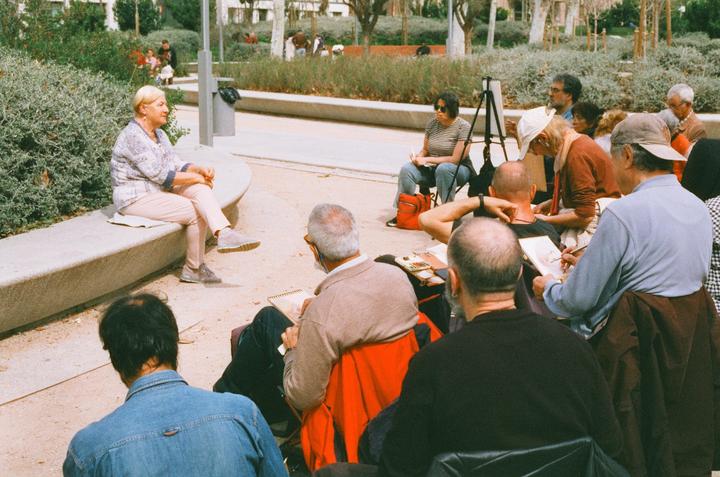 A group of people are drawing a woman sitting on a concrete bench in a park.