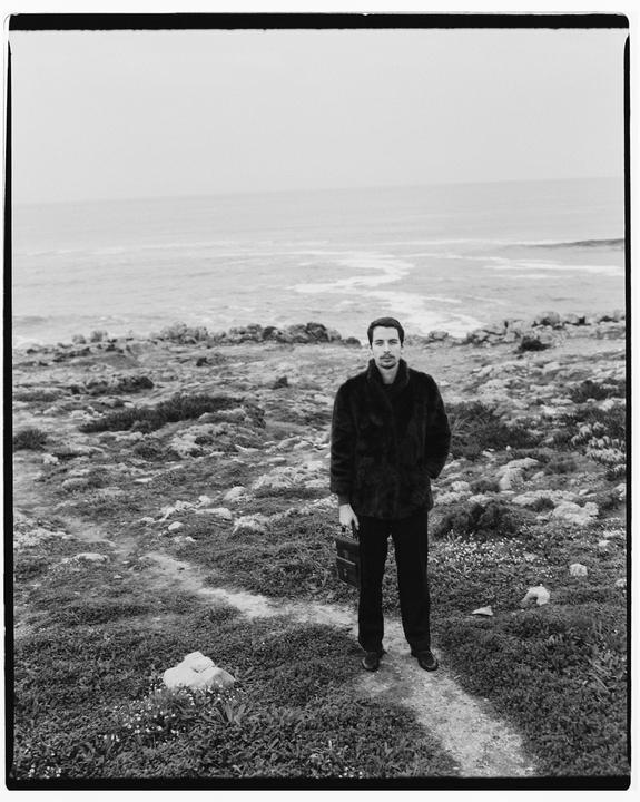 A man in a fur coat stands on a path overlooking the ocean.