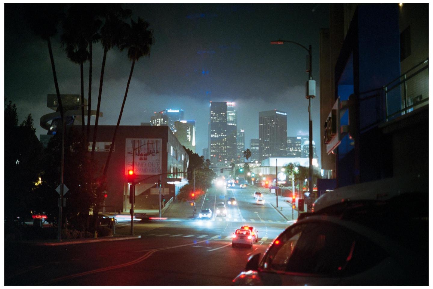 A street scene at night shows cars driving towards a city skyline with palm trees on the left.