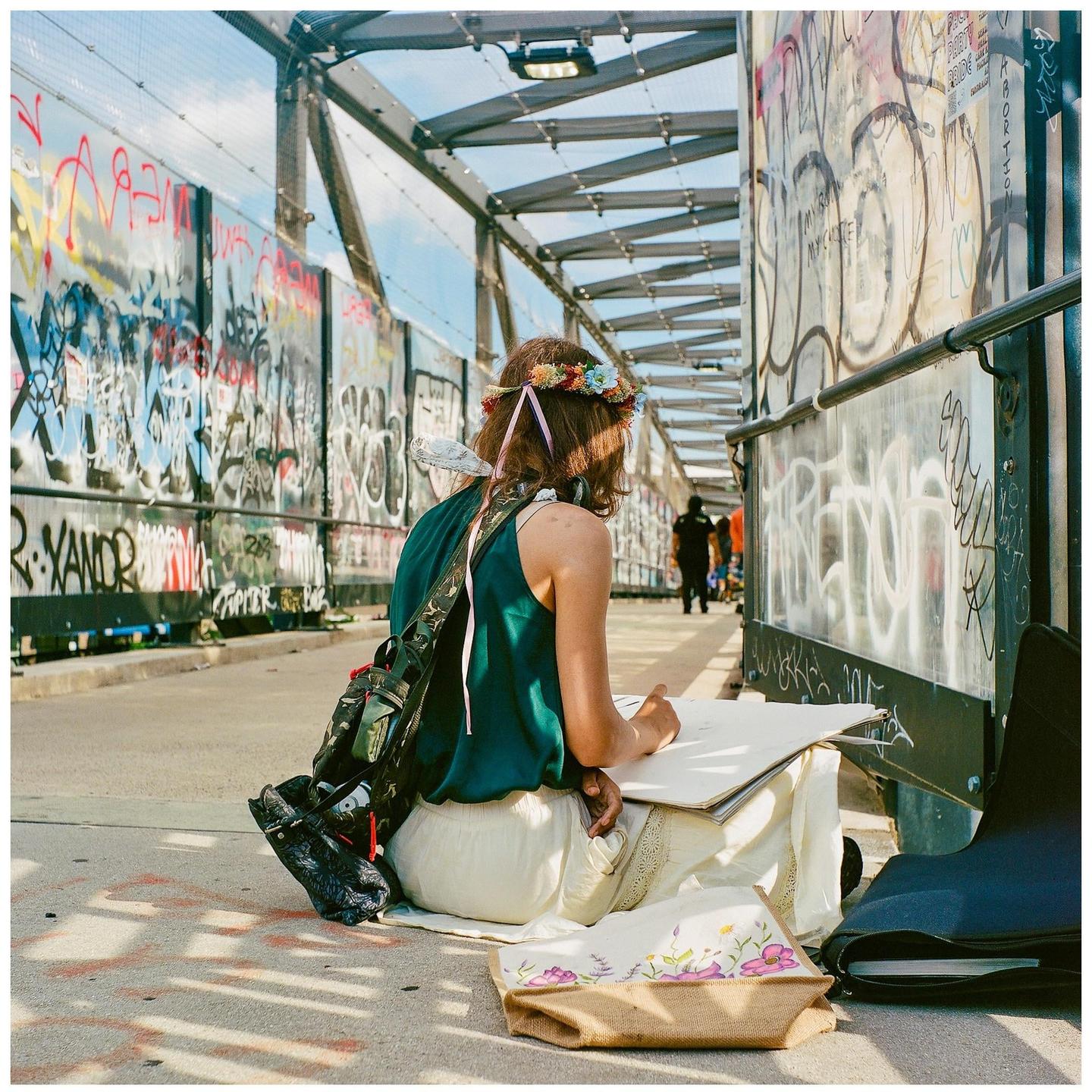 A woman wearing a flower crown sits on a bridge covered in graffiti and draws in a sketchbook.