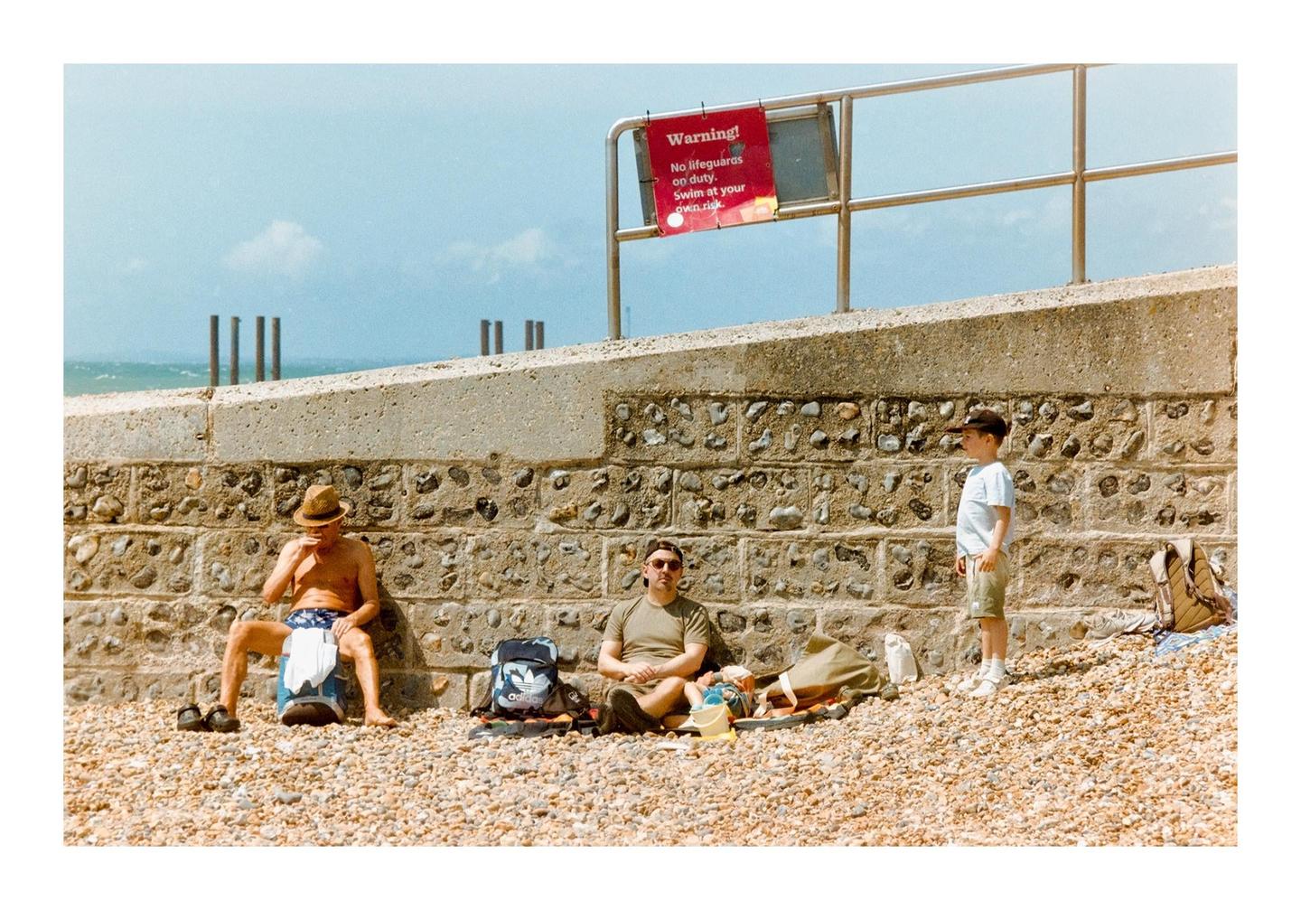Three people are relaxing on a pebble beach next to a stone wall with a warning sign above it.