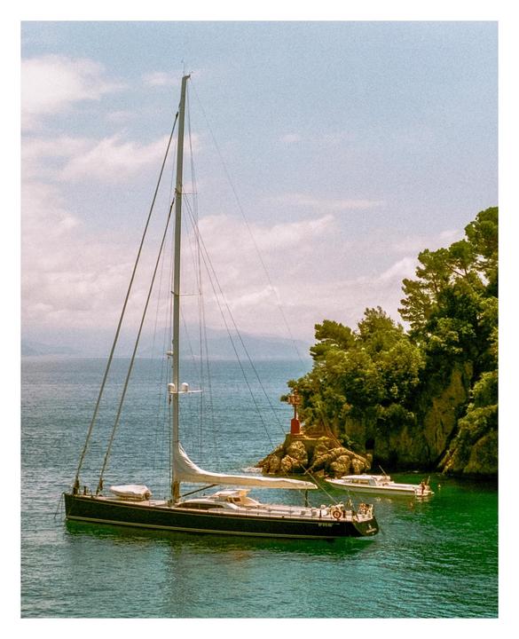 A sailboat and a motorboat are anchored in a cove with a small lighthouse on a rocky, tree-covered shore.