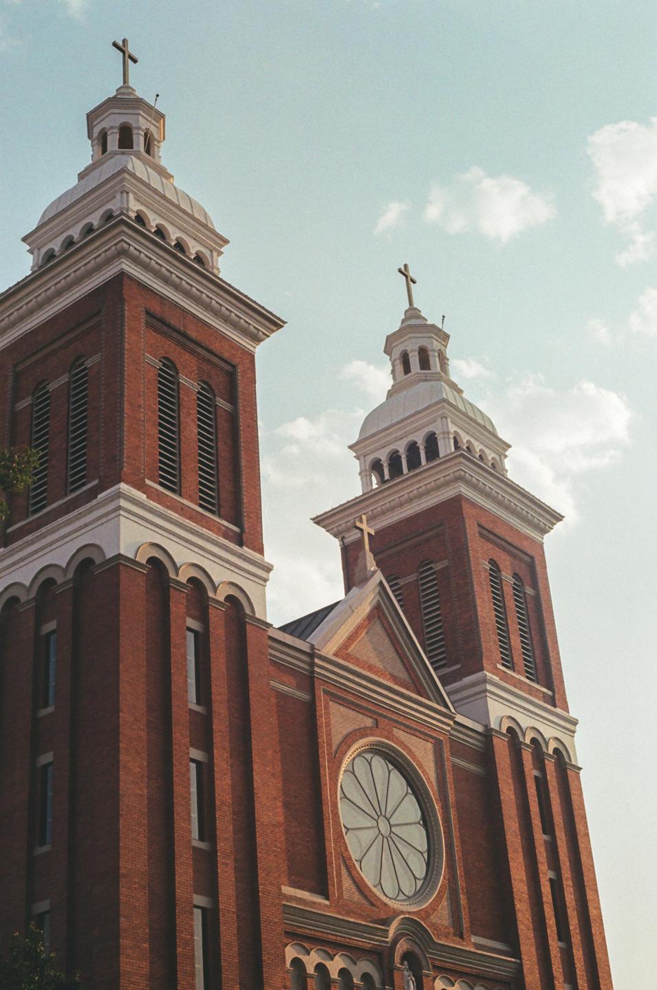 A brick church with two towers and a circular window is shown from a low angle against a cloudy sky.
