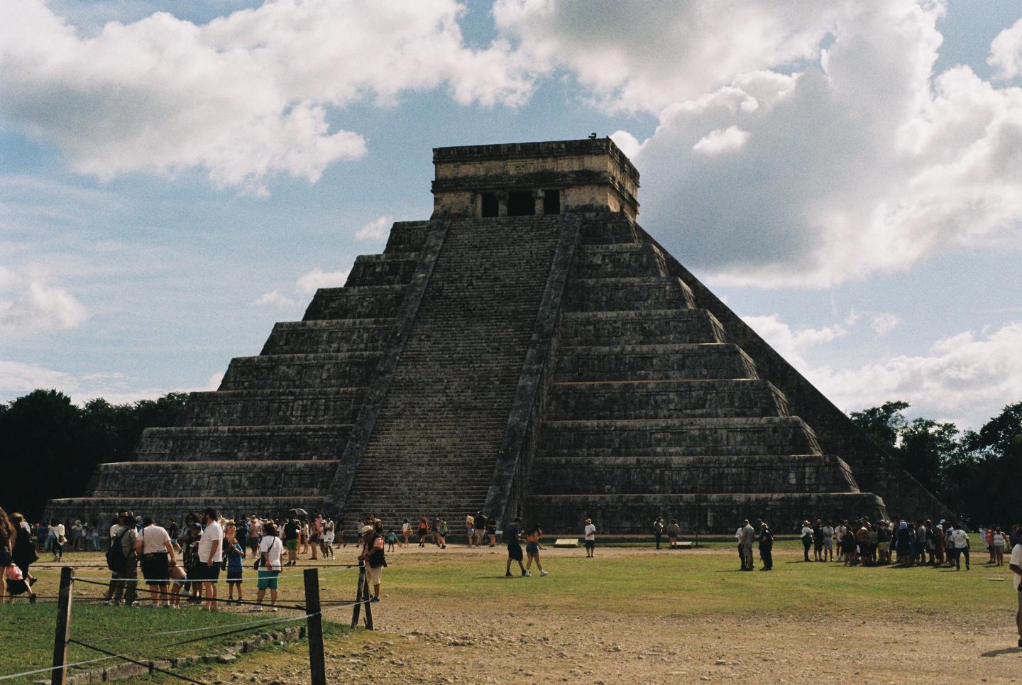 A large stone pyramid with many steps stands under a cloudy sky, surrounded by people on a grassy field.