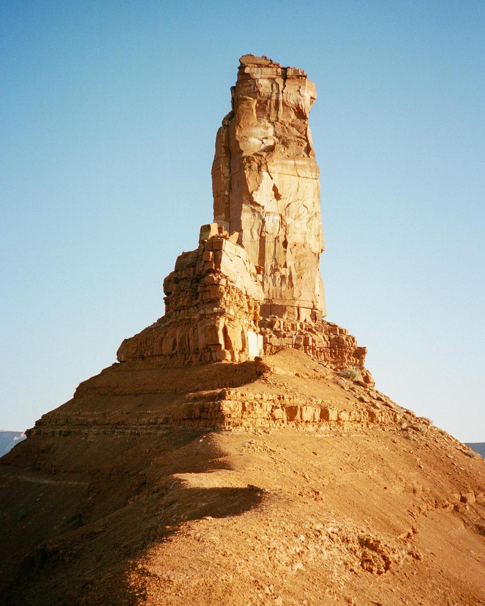 A tall, narrow sandstone formation rises from a desert landscape under a clear sky.
