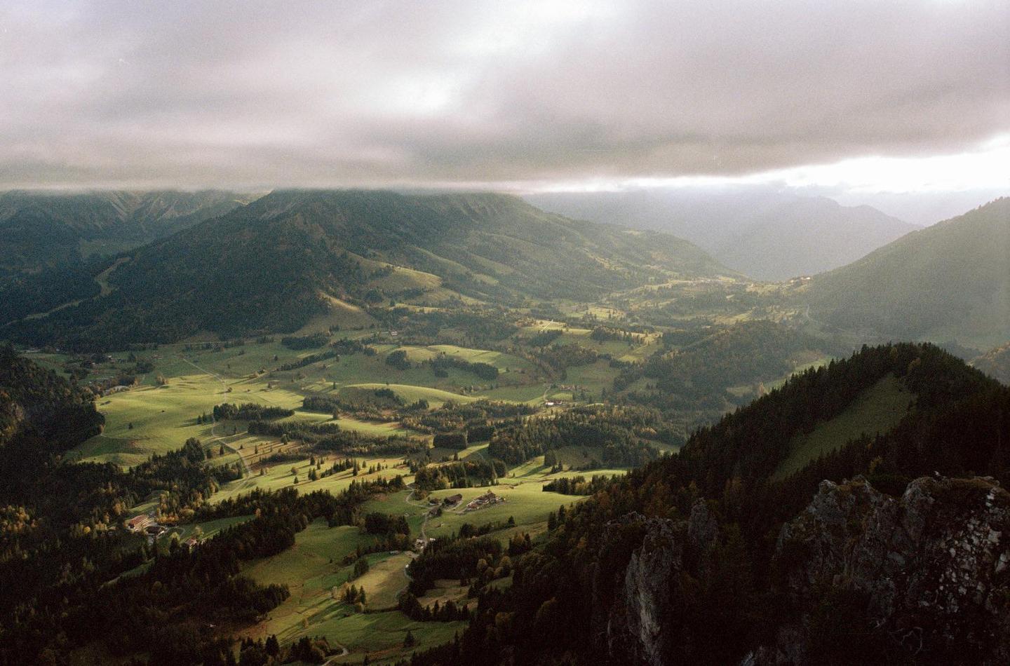 A wide valley with green fields and scattered trees is nestled between mountains under a cloudy sky.