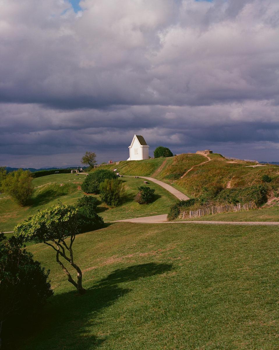 A small white building sits atop a grassy hill under a cloudy sky, with winding paths and trees in the foreground.