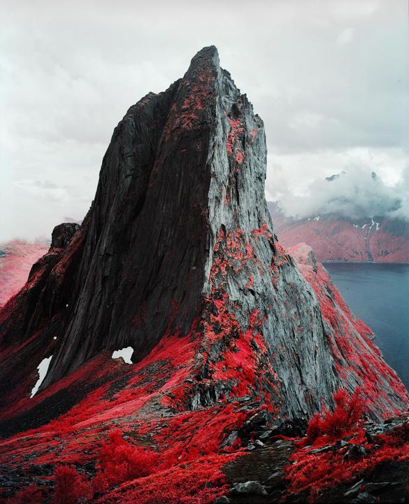 A large, jagged mountain with vibrant red foliage at its base rises above a body of water under a cloudy sky.