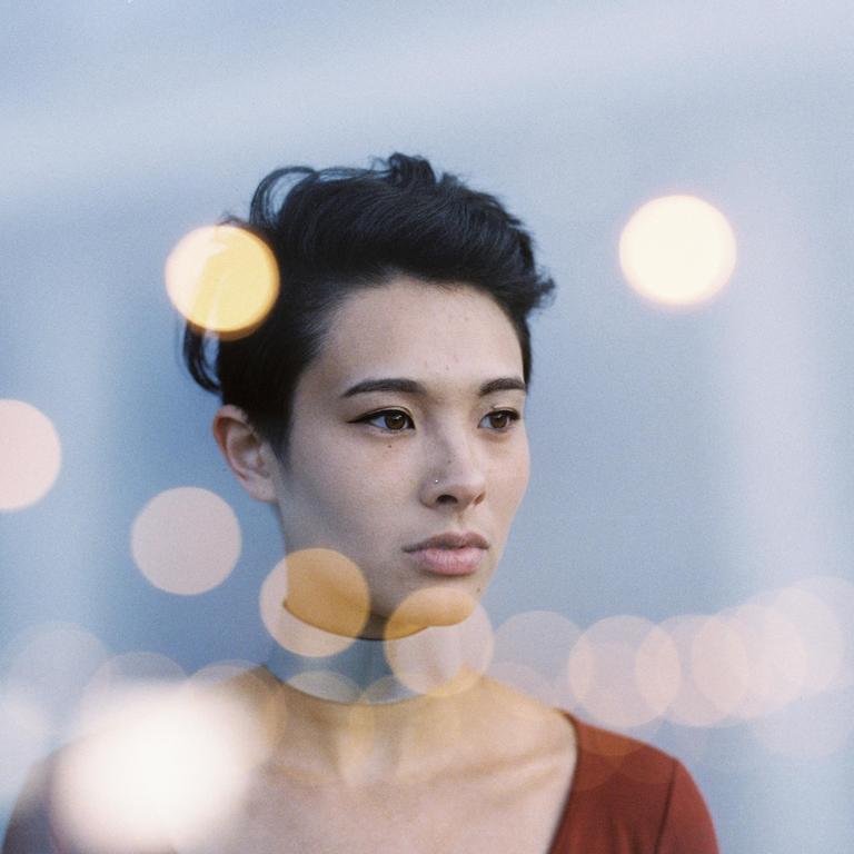 A woman with short dark hair and a nose piercing looks to the side, with bokeh lights in the foreground and background.