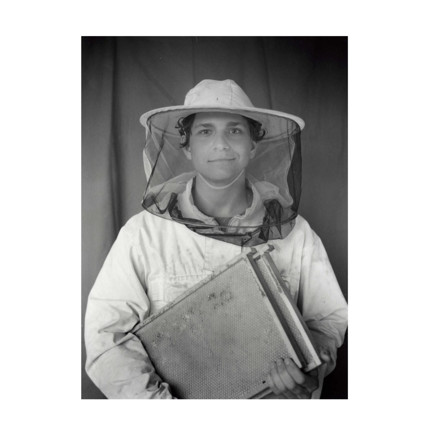 A young man wearing a beekeeper suit and hat holds a honeycomb frame.