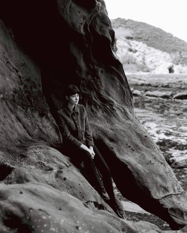 A woman sits on a large rock formation with a rocky landscape and hill in the background.