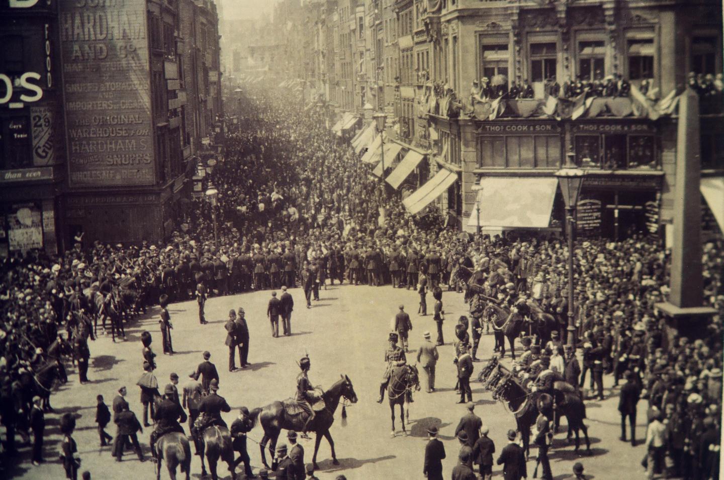 A large crowd of people and soldiers on horseback fill a city street lined with buildings.