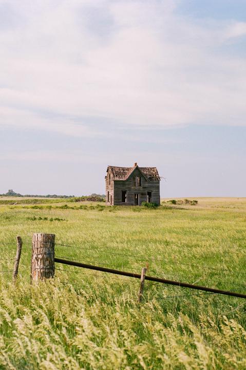 An abandoned wooden house with a damaged roof stands in a field of tall grass under a cloudy sky, with a barbed wire fence in the foreground.
