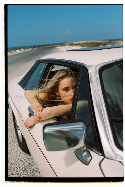 A woman with long blonde hair leans out of a car window on a sunny day.