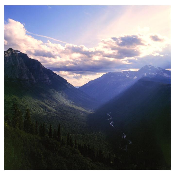A valley with a river and mountains is illuminated by beams of light from a cloudy sky.