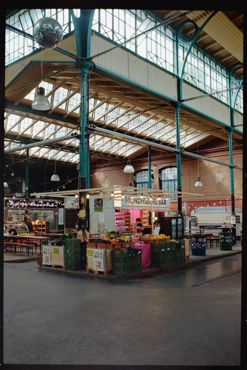 A disco ball hangs from the ceiling of a market hall with various stalls and people.
