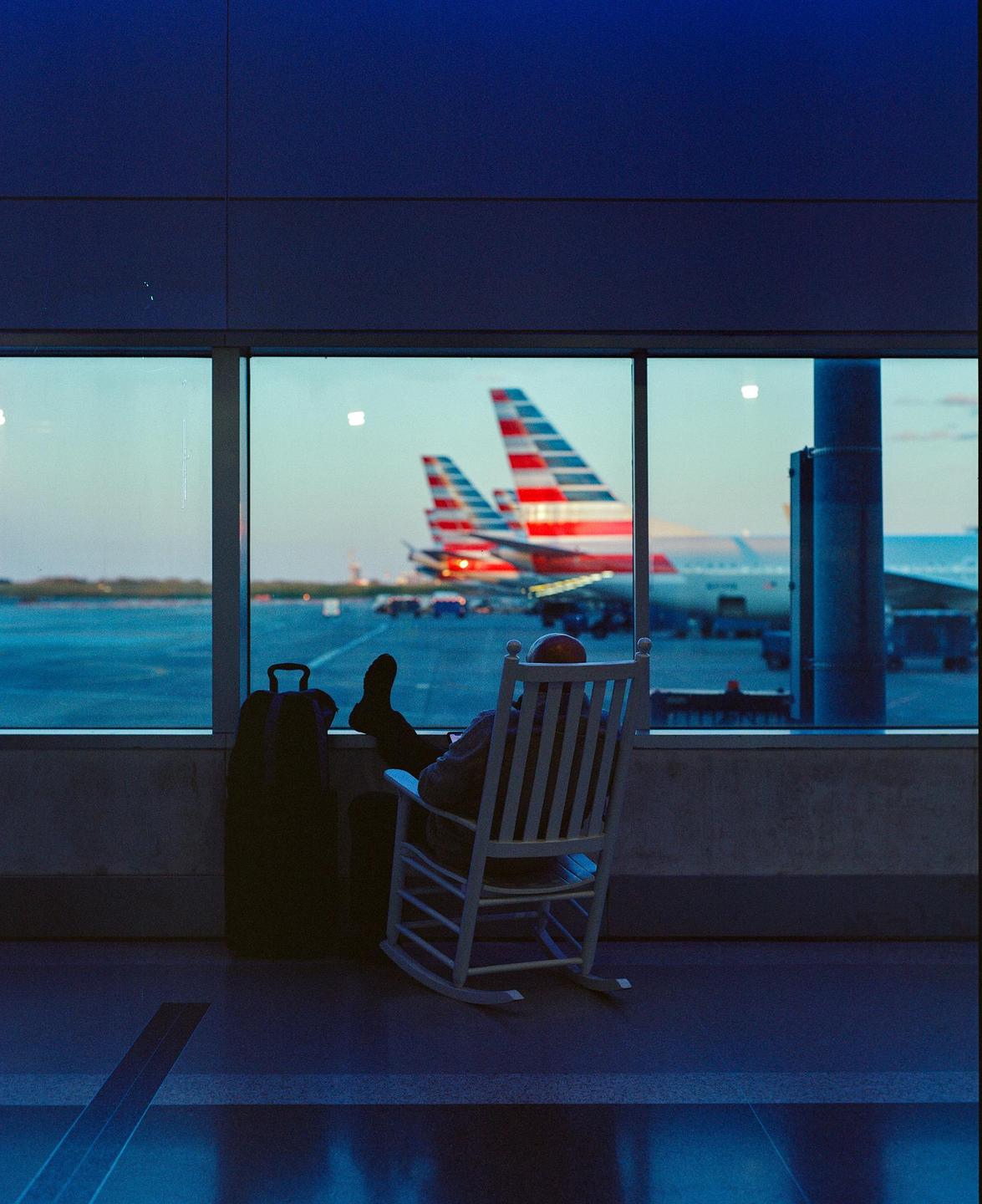 A person sits in a rocking chair with their feet up, looking out an airport window at two airplanes.