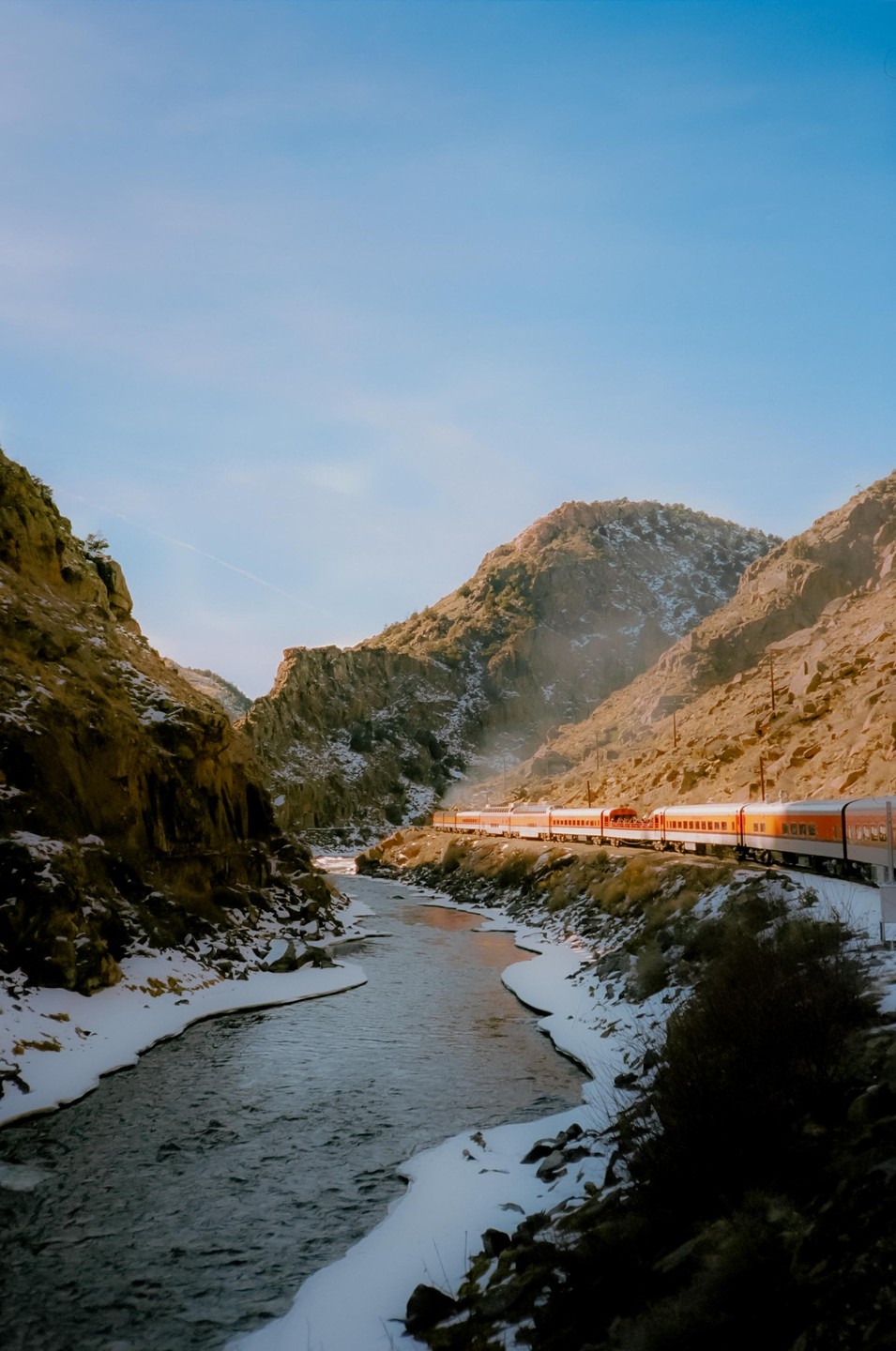 A train travels along a river through a snowy mountain canyon.