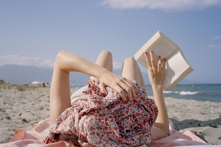 A woman lies on a towel on a sandy beach, reading a book, with the ocean and mountains in the background.
