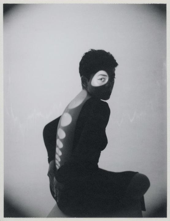 A woman with short hair sits with her back to the viewer, looking over her shoulder, with light projected onto her face and back.
