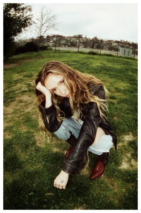 A woman with long hair, wearing a leather jacket, ripped jeans, and cowboy boots, crouches in a grassy field.