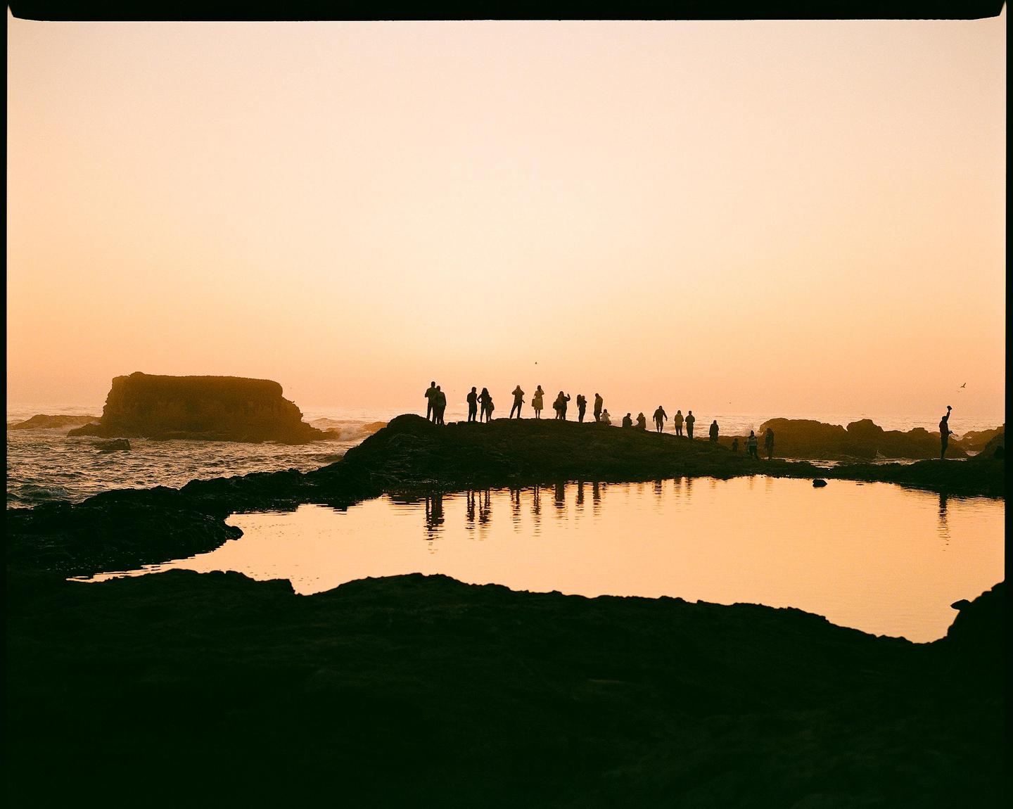 A group of people stand on a rocky outcrop overlooking the ocean at sunset, with their reflections visible in a tidal pool.