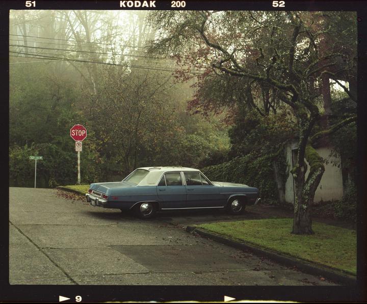 A blue car is parked on a street corner next to a stop sign on a foggy morning.