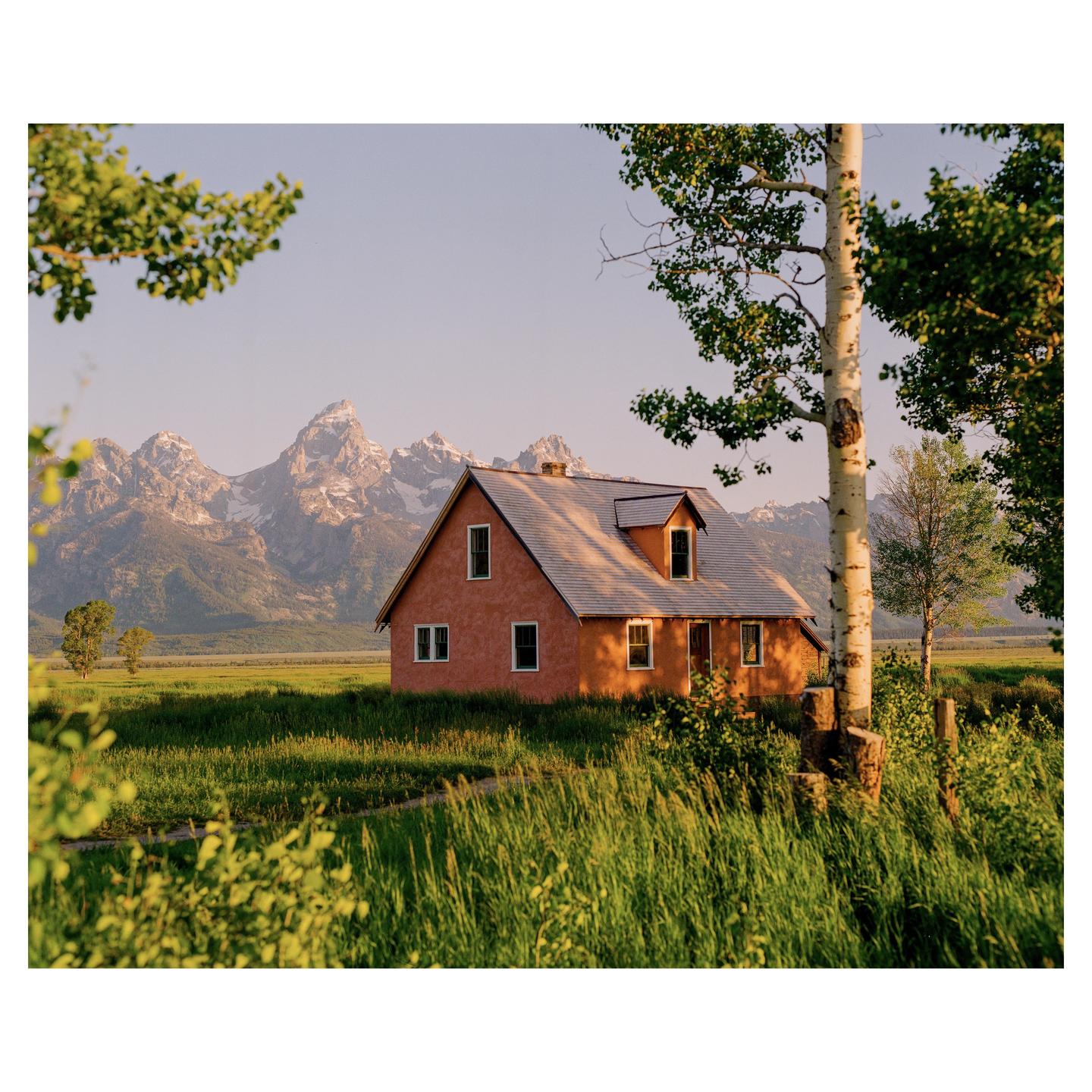 A red house stands in a field of tall grass with snow-capped mountains in the background.