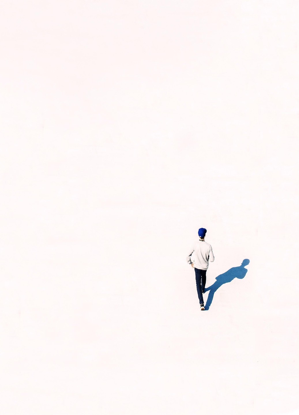 A man in a blue hat walks across a bright white surface, casting a long blue shadow.