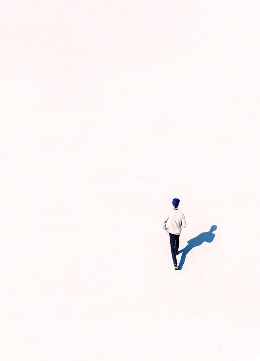 A man in a blue hat walks across a bright white surface, casting a long blue shadow.