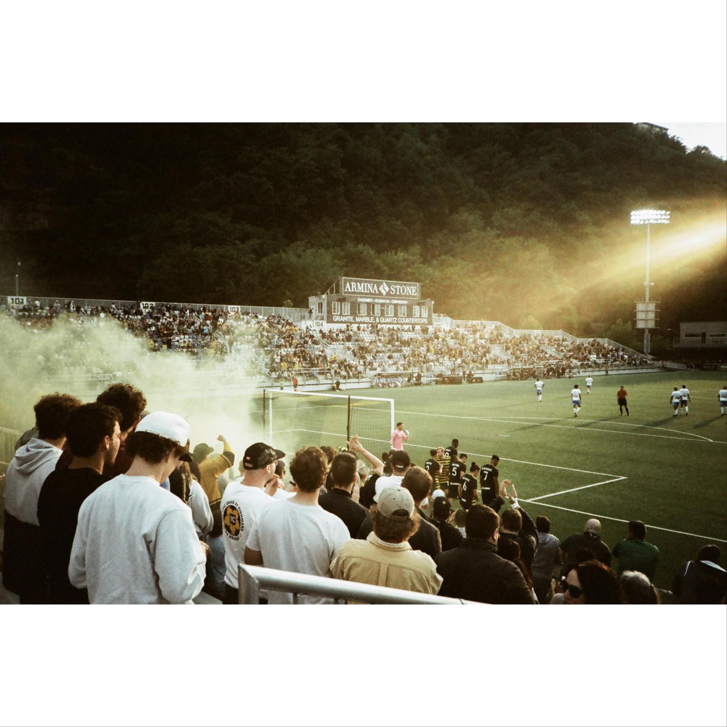 A crowd of people watches a soccer game in a stadium with a hillside in the background.