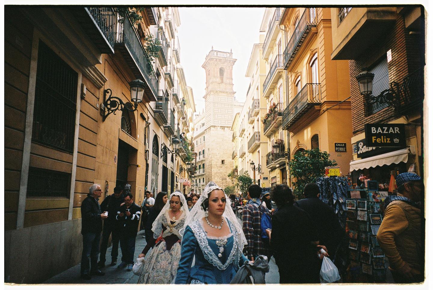 Two women in traditional dresses and lace veils walk down a narrow street lined with buildings and people, with a tall tower visible in the distance.
