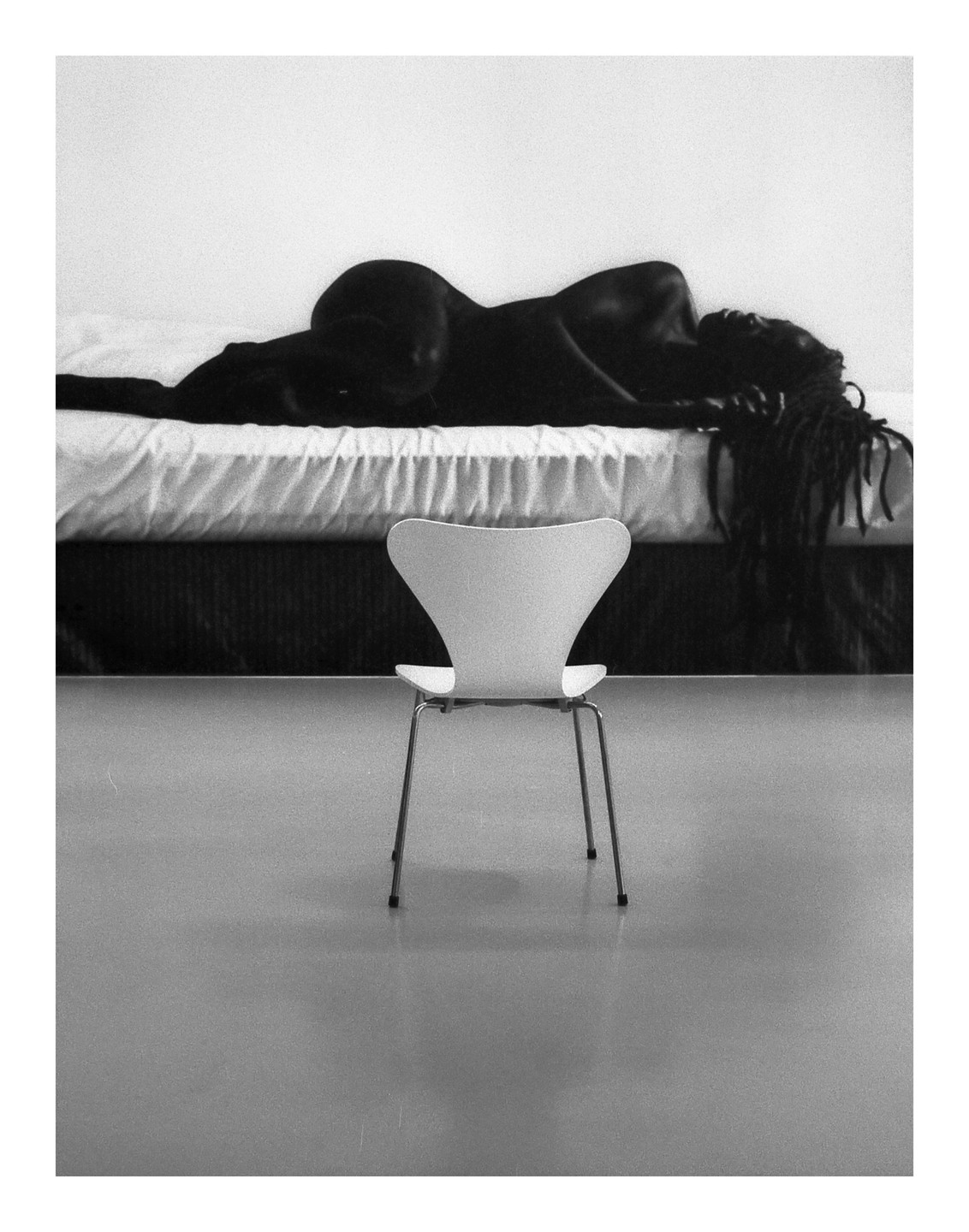 A woman with dreadlocks lies on a bed in a room with a white chair in the foreground.