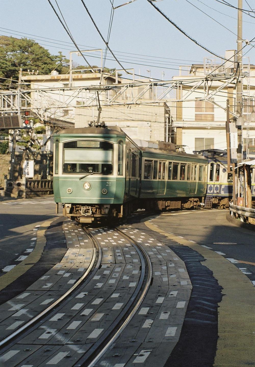 A green tram travels along a curved track in a city with buildings and power lines in the background.