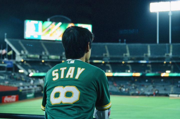 A person wearing a green jersey with "STAY 8" on the back watches a baseball game at night.