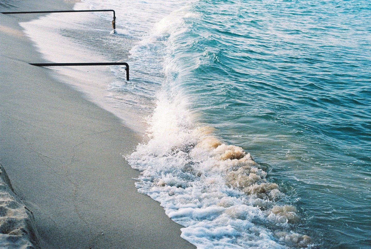Waves crash on a sandy beach with two metal pipes extending into the water.