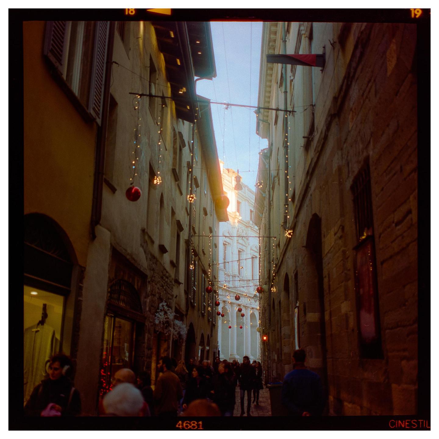 A narrow street in an old town is decorated with hanging lights and red ornaments, with people walking below.