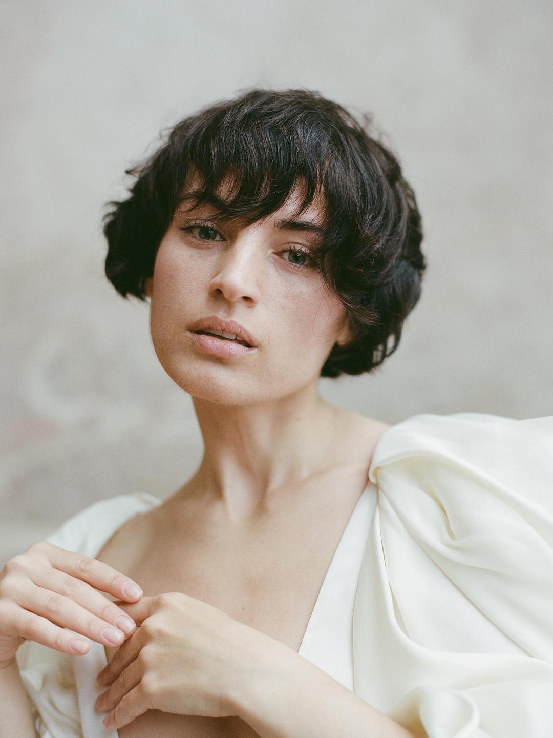 A woman with short dark hair and a white garment looks directly at the viewer.
