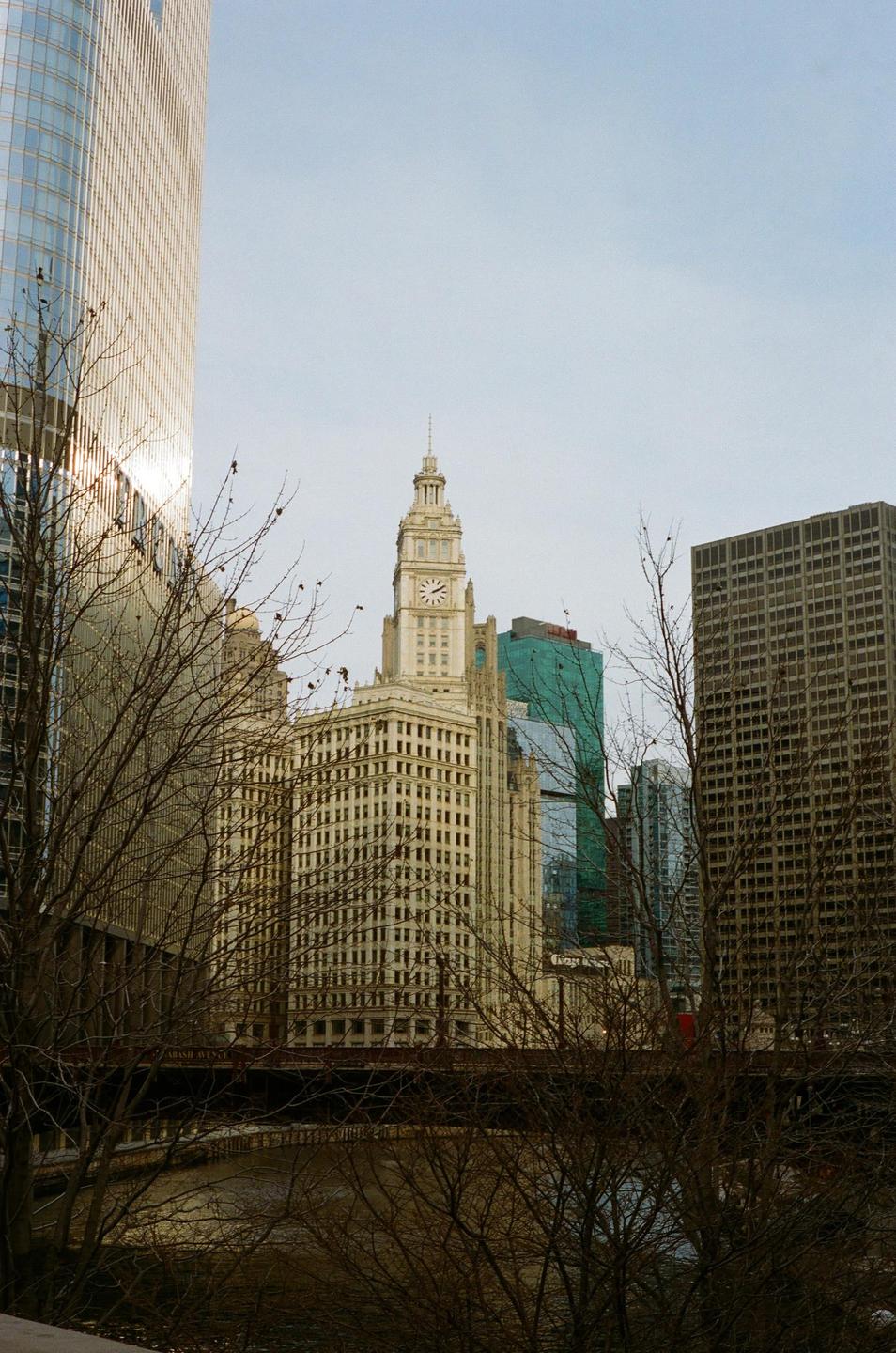 A clock tower rises above other buildings and bare trees next to a river.