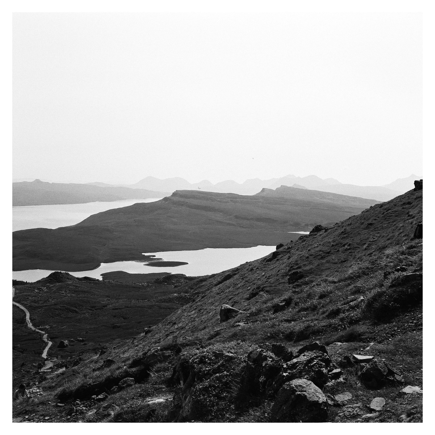 A winding path leads down a steep, grassy hill towards a body of water with mountains in the distance.