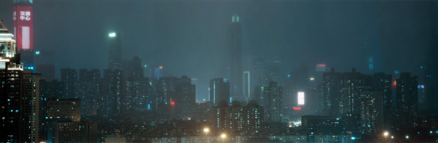 A panoramic view of the Shenzhen skyline at night shows numerous illuminated buildings and skyscrapers shrouded in a hazy atmosphere.